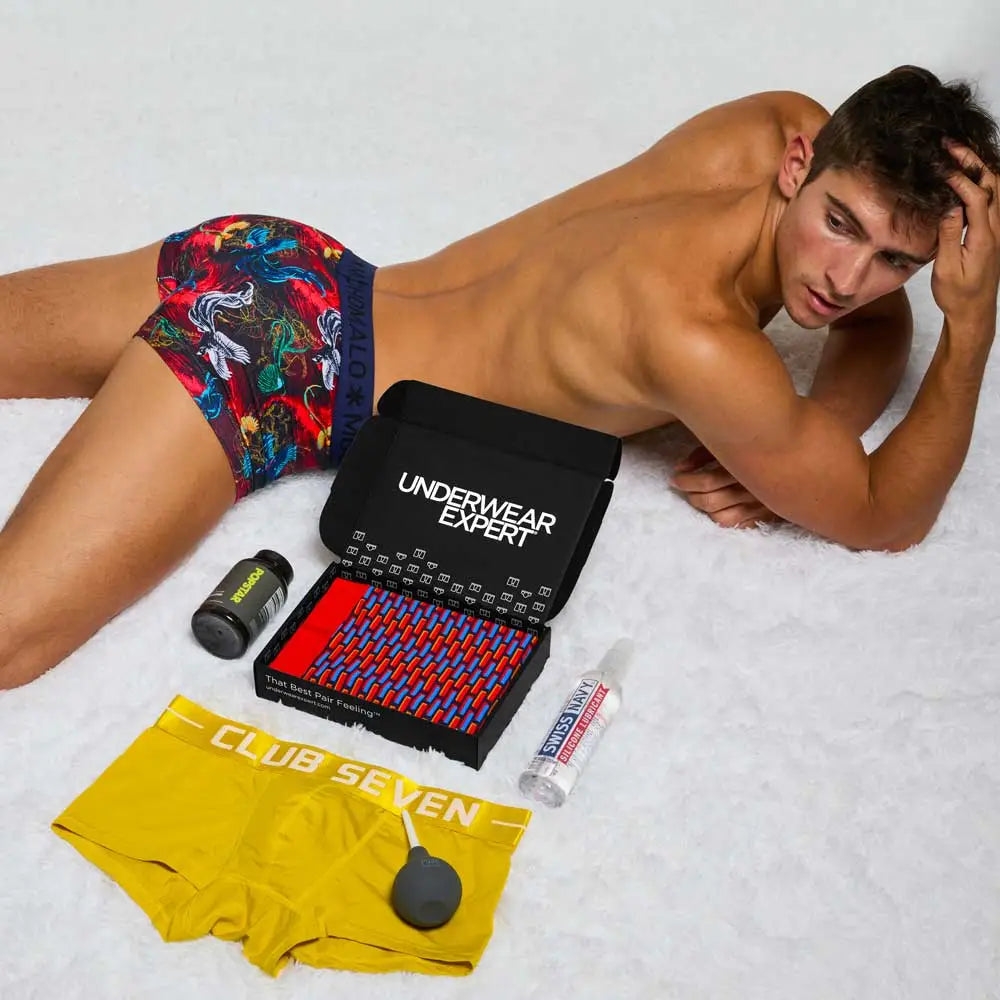 Man lying on a bed with colorful underwear and 'Underwear Expert' packaging.